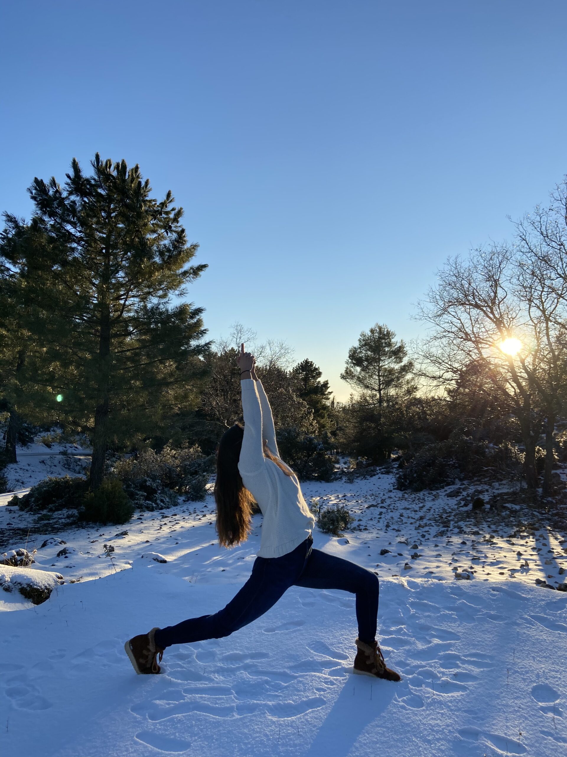 Mujer con ropa de invierno haciendo la postura del guerrero numero uno sobre nieve y con arboles al fondo.